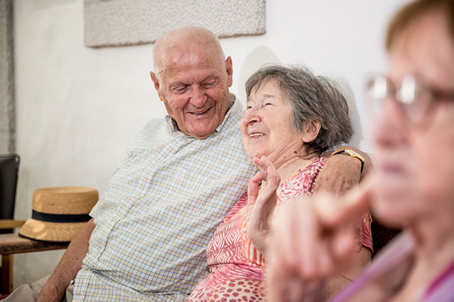 An elderly man and an elderly woman sitting on a couch, his arm is around the woman, he is smiling at her and she is laughing